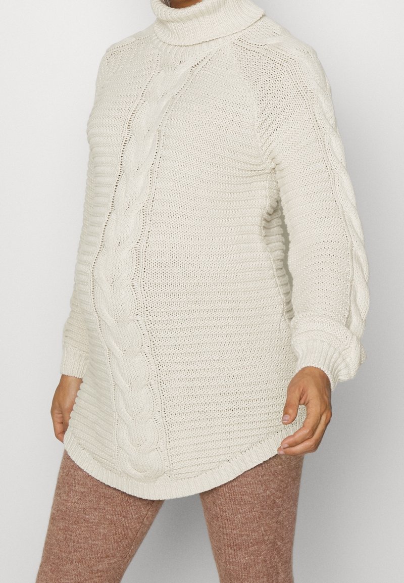 Pull à col roulé en maille torsadée de couleur crème avec manches longues, porté avec un pantalon en maille marron clair, visible du cou jusqu'à mi-cuisse.