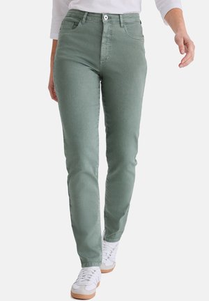 Frau, die hellgrüne, figurbetonte Jeans, weiße Sneaker und ein weißes Langarmshirt trägt und vor einem einfarbigen Hintergrund läuft.