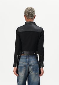 Giacca nera a maniche corte con dettagli in pelle, caratterizzata da un colletto e zip sulle maniche, abbinata a jeans in denim blu con cuciture distintive.
