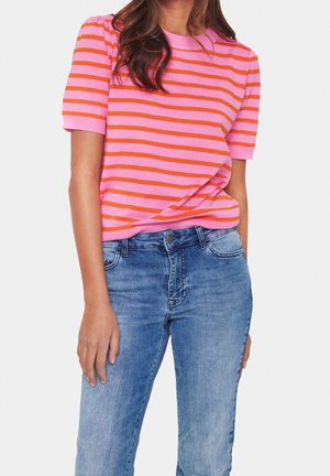 Vrouw draagt een kortärmelig top met roze en oranje horizontale strepen, gecombineerd met lichtblauwe spijkerbroek, staand tegen een witte achtergrond.