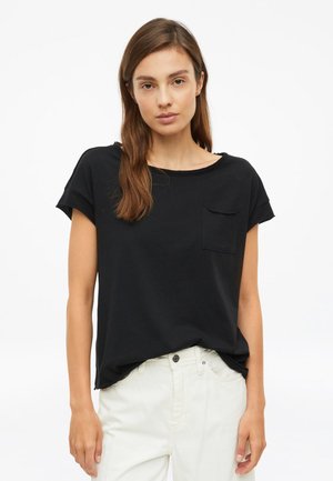 Frau mit langen braunen Haaren, die ein schwarzes Kurzarmshirt mit Fronttasche und eine weiße Hose trägt, steht vor einem schlichten weißen Hintergrund.