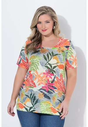 Kvinde med blondt hår iført sølv øreringe og en farverig kortærmet skjorte med tropiske blomsterprint, stående mod en hvid baggrund.