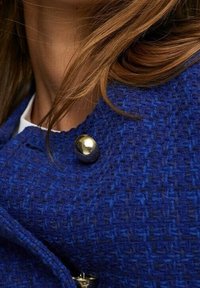 Veste textile bleue tissée présentant un grand bouton métallique doré et un motif texturé, avec un gros plan sur la zone du col.