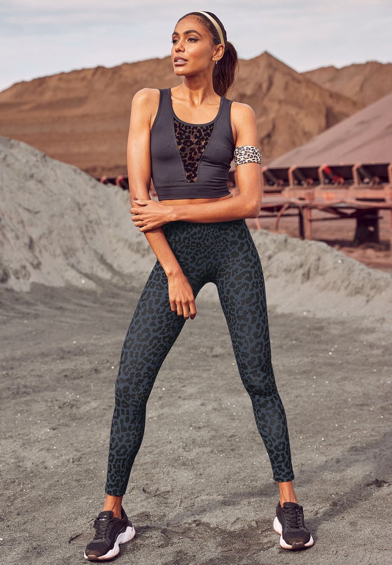 Zwarte sportbeha met een lederen inzet, gecombineerd met high-waisted leggings met een zwart luipaardpatroon. Zwarte sportschoenen.