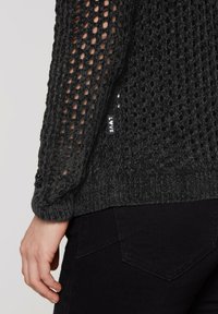 Schwarzer offener Strickpullover mit langen Ärmeln, aus einem strukturierten Stoff und mit einem kleinen "LOVE"-Etikett am Saum. Über einer schwarzen Hose getragen.