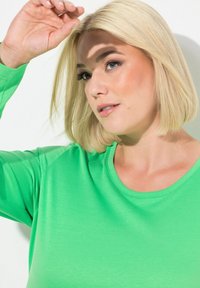 Helles grünes Langarmshirt mit rundem Ausschnitt und weicher Textur. Die Haare sind im Bob geschnitten und verdecken teilweise das Gesicht, während eine Hand erhoben ist.