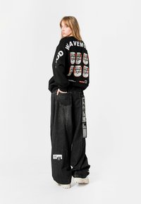 Sudadera negra con estampado gráfico y texto en la parte posterior. Jeans grises de gran tamaño con etiquetas. El modelo lleva zapatos de color claro. Fondo blanco.