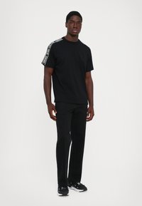 Άνδρας με μαύρη μπλούζα Versace με λογότυπο στους ώμους, μαύρο παντελόνι και μαύρα αθλητικά παπούτσια, που στέκεται μπροστά σε λευκό φόντο.