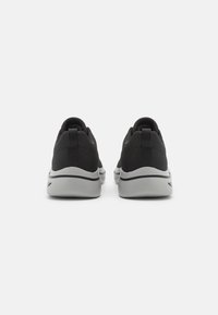 Scarpe da ginnastica nere con una parte superiore texturizzata, forma arrotondata e suole in gomma grigia. Le caratteristiche includono una linguetta posteriore e linee di design sottili.