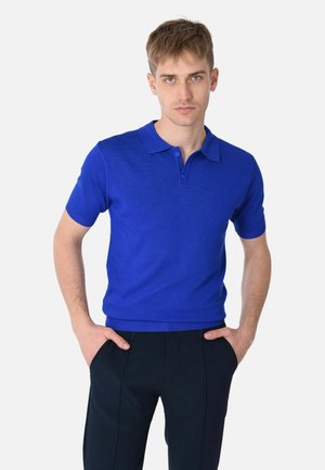 Giovane uomo con capelli corti in piedi con le mani nelle tasche, che indossa una polo a maniche corte di un azzurro brillante e pantaloni scuri su uno sfondo bianco.