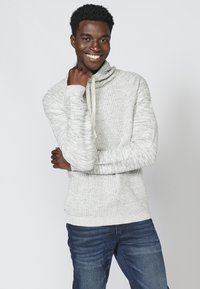 Hellgrauer gestrickter Pullover mit einem Schalkragen und einem Kordelzug, kombiniert mit dunkelblauen Jeans. Die Textur ist grob und weich.