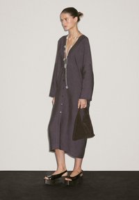 Frau steht in einem dunklen, übergroßen, knopfartigen Kleid, trägt eine lange Halskette, hält eine kleine dunkle Handtasche und trägt schwarze Plateausandalen vor einer schlichten Wand.