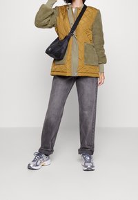 Person i en senapsgul quiltad väst med gröna fleeceärmar, grå jeans, silverfärgade sneakers och en svart crossbody-väska mot en enfärgad bakgrund.