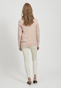 Ljusrosa tröja med ribbad mudd och nederkant, tillsammans med krämfärgade åtsittande jeans och svarta klackskor. Håret är långt och vågigt.