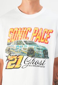 T-shirt en coton blanc avec un graphique multicolore d'une voiture de course et un texte en gras "SONIC PACE" et "21 Ghost Worldwide", avec un effet vieilli.