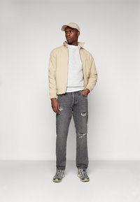 Chaqueta bomber beige sobre un sudadera gris, jeans rasgados grises y zapatillas multicolores. Una gorra beige clara completa el atuendo.