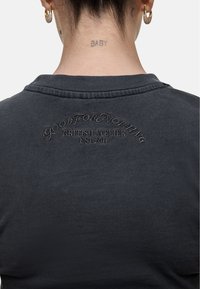 Camicia in cotone grigio scuro con colletto, testo ricamato sul retro e una texture liscia con dettagli minimi.