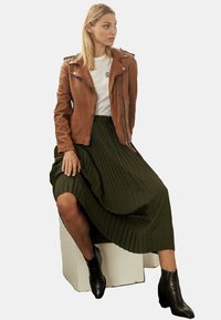 Braune Lederjacke, weißes T-Shirt mit Print, olivgrünem gefälteltem Midikleid und schwarzen Ankle Boots mit strukturiertem Finish.