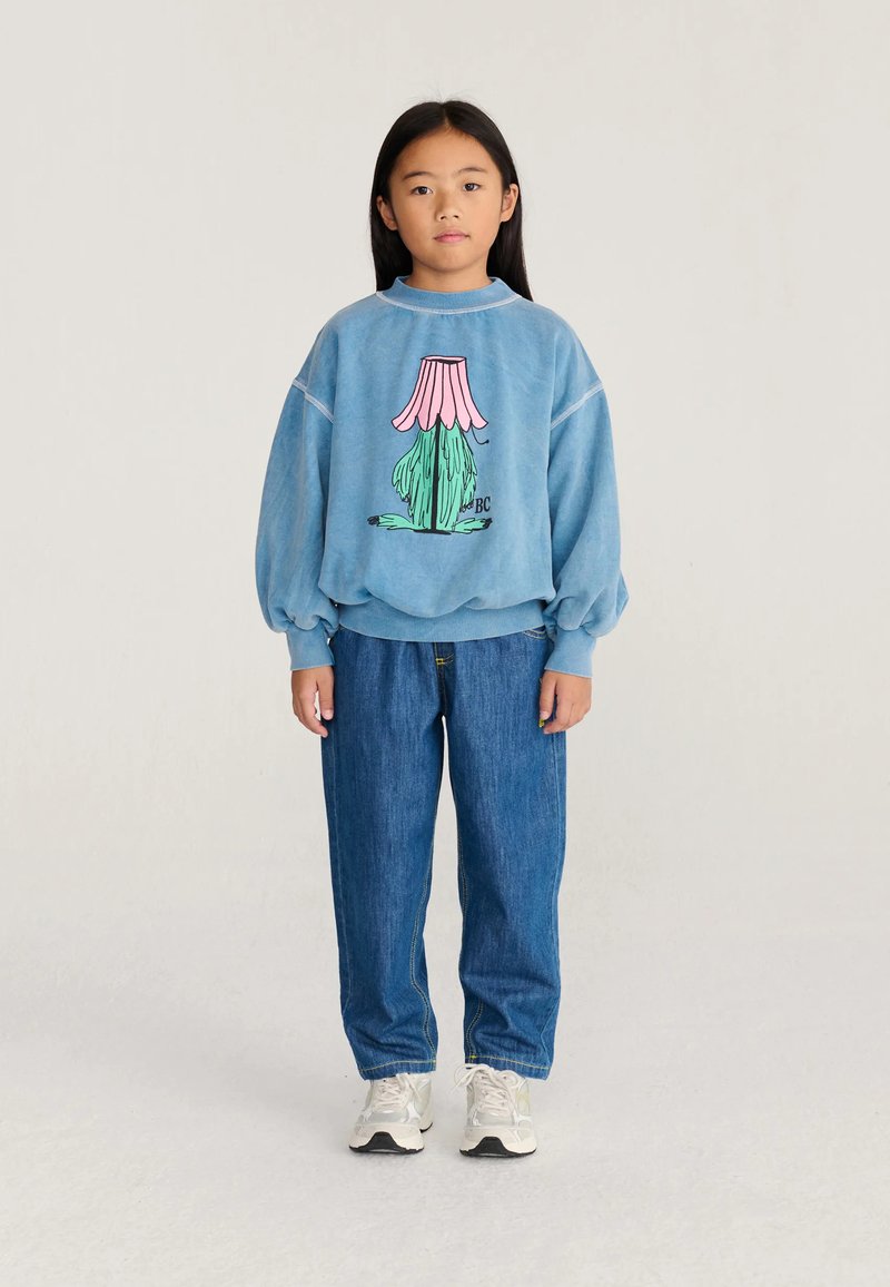 Jeune fille debout portant un sweat-shirt bleu avec un graphisme fleuri coloré, un jean bleu et des baskets blanches sur un fond uni.