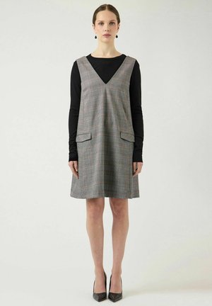 Femme portant une robe sans manches à carreaux longueur genou sur un haut noir à manches longues, associée à des escarpins noirs, debout devant un fond uni.