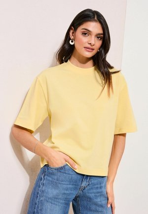 Ung kvinde med mørkt hår iført hvide hoops øreringe, en lysegul kortærmet skjorte og blå jeans, der står op ad en lys væg.