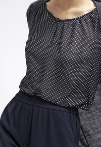 Svartvit prickig blus med rynkad halsringning, parat med ett marinblått elastiskt midjeband, under en kolgrå cardigan.