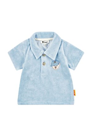 Light blue velour polo shirt with collar, three buttons, and an embroidered bear patch wearing a striped cap. Small brand label on side.