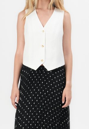 Vrouw draagt een mouwloos wit vest met knopen, gecombineerd met een zwarte geplooide rok met witte stippen.