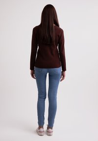 Brauner langärmeliger Pullover kombiniert mit hellblauen Skinny-Jeans und weißen Sneakers mit rosa Akzenten. Modell von hinten betrachtet.