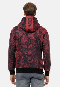Roter und schwarzer Hoodie mit grafischem Löwen-Design auf dem Rücken, ausgestattet mit einer Kapuze mit Kordelzug und gerippten Bündchen. Glatte Stoffoberfläche.