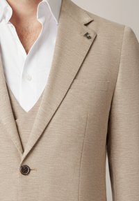 Mężczyzna w beżowej, teksturowanej marynarce i kamizelce noszący białą koszulę z otwartym kołnierzykiem.