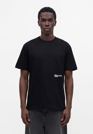 Ung mand med kort krøllet hår iført en sort Karl Lagerfeld Jeans t-shirt og mørke jeans mod en ensfarvet hvid baggrund.