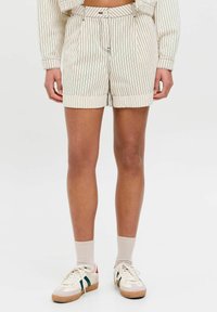 Pantaloni corti color crema con righe verticali nere, dotati di chiusura a bottone, risvolti e tasche frontali. Indossati con sneakers e calzini.
