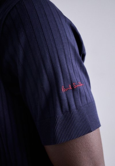 Haut à manches courtes côtelé bleu marine avec une coupe ajustée et un ourlet texturé avec de larges côtes. Un petit logo rouge "Paul Smith" est présent sur la manche.