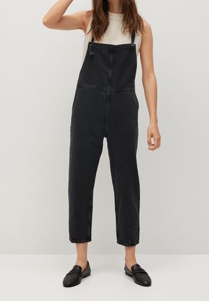 Nainen, joka pukeutunut valkoiseen hihattomaan toppiin, mustiin farkkuhaalarikuoseihin ja mustiin slip-on-kenkiin, tasaisen taustan edessä.