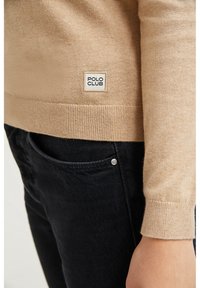 Beige knit sweater with a ribbed hem and cuff, featuring a white logo patch labeled "POLO CLUB." Paired with black jeans showing a pocket detail.
