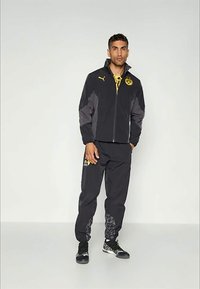 Mann iført svart og grå Puma treningsdrakt med gule Borussia Dortmund-logoer, som står mot en ensfarget lys grå bakgrunn.