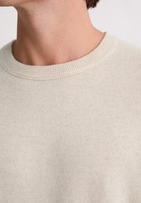 Jasno beżowy sweter z prążkowanym okrągłym dekoltem, miękką fakturą i swobodnym krojem. Zbliżenie na obszar szyi i detale kołnierza.