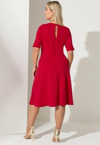Czerwona sukienka z krótkimi rękawami, fason w kształcie litery A, detal z otworem w tylnej części oraz zamek błyskawiczny. Noszona z złotymi sandałkami na obcasie. Gładka faktura materiału.