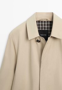 Beige trenchcoat met spitse kraag, één zichtbare zwarte knoop en een geruite voering met label "Massimo Dutti."