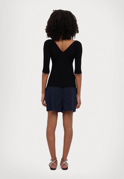 Haut noir à côtes, ajusté, avec un décolleté en V et des manches trois-quarts, associé à un short marine. Les chaussures sont des sandales à lanières avec une semelle plate.