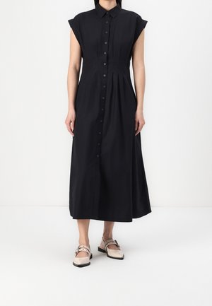 Femme portant une robe noire midi sans manches à boutons avec col, et des chaussures plates beiges avec des brides et des boucles.