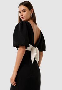Abito nero con maniche a sbuffo, schiena scoperta e un nastro bianco legato in vita. Il tessuto strutturato esalta il design dell'abito.