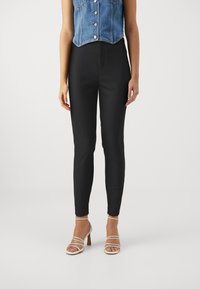 Sorte, slim-fit bukser med en glat tekstur; har høj talje og ingen synlige knapper eller lynlåse, kombineret med stroppede beige hæle.