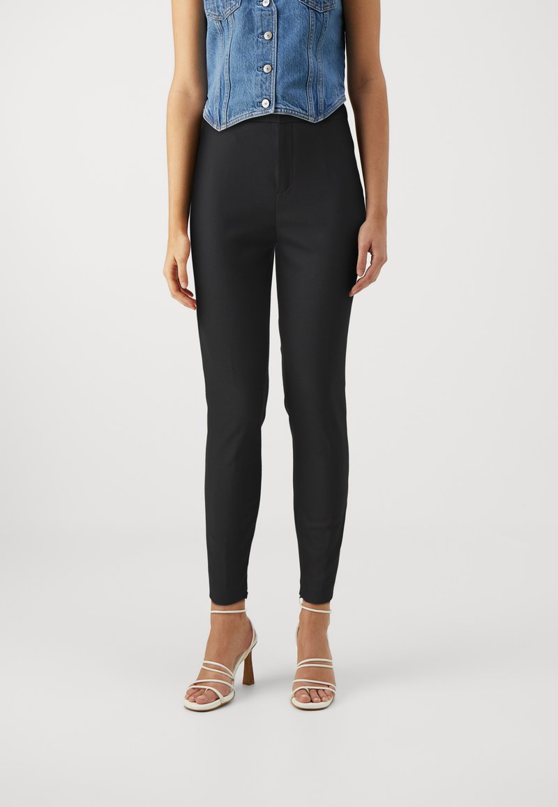 Sorte, slim-fit bukser med en glat tekstur; har høj talje og ingen synlige knapper eller lynlåse, kombineret med stroppede beige hæle.