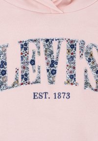 Rožinė striukė su "LEVI'S" gėlių rašto raidėmis, po kuriais mėlynu užrašyta "EST. 1873". Minkštas audinio tekstūra.