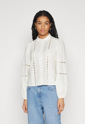 White blouse with long puff sleeves, crochet detailing along button placket and sleeves, and a cropped design. Paired with blue jeans.