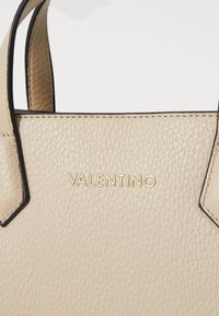 Bežinė odinė rankinė su tekstūruota paviršiumi, su juodu apvadu ir auksiniu "Valentino" prekės ženklu priekyje. Rankenos viršuje.
