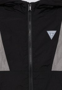 Veste noire avec fermeture éclair, comportant un panneau gris et un logo triangulaire Guess sur la poitrine. Design simple et sportif.