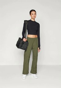 Kvinne som står iført en svart langermet crop top, olivengrønne bukser, hvite sneakers og bærer en svart skulderveske.